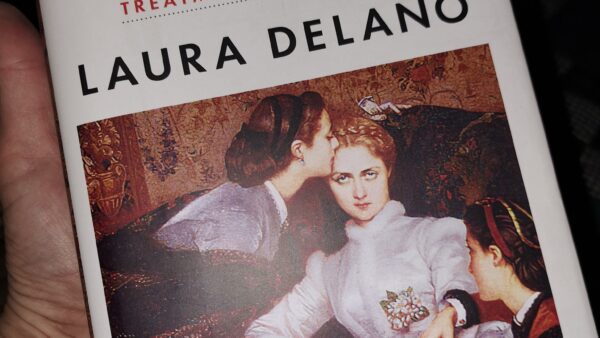 A hand holds the book Unshrunk: A Story of Psychiatric Treatment Resistance by Laura Delano. The cover features a classical painting of three women in period clothing, one comforting another with a kiss on the temple. A quote at the bottom reads, "A must read." —Anna Lembke, MD, author of Dopamine Nation.